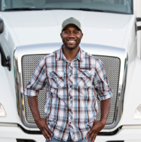 Man truck driver wearing a cap, standing in front of his white freightline with his hands in his pockets, smiling at the camera. 